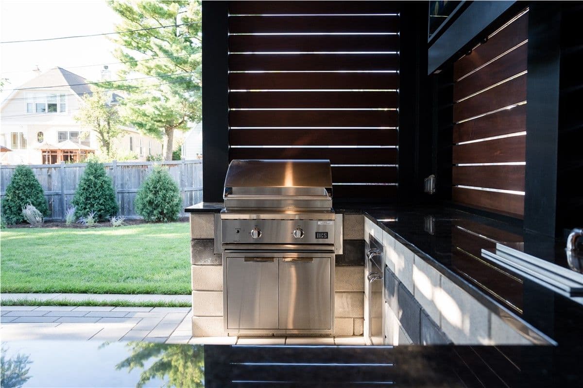A stone outdoor grilling station and kitchen with stainless steel grill underneath a pergola looking out onto a backyard.