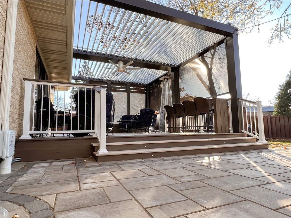 A brown deck with white fencing and a black pergola with a sun screen and privacy screen. Dark patio furniture and bar stools sit on the deck.
