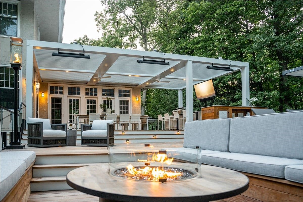 Outdoor kitchen and dining area with a sunken stone fire pit and seating, built-in stainless steel grill, bar seating with whitestools, and a white pergola, set on a multi-level wood deck with surrounding trees and landscaping.