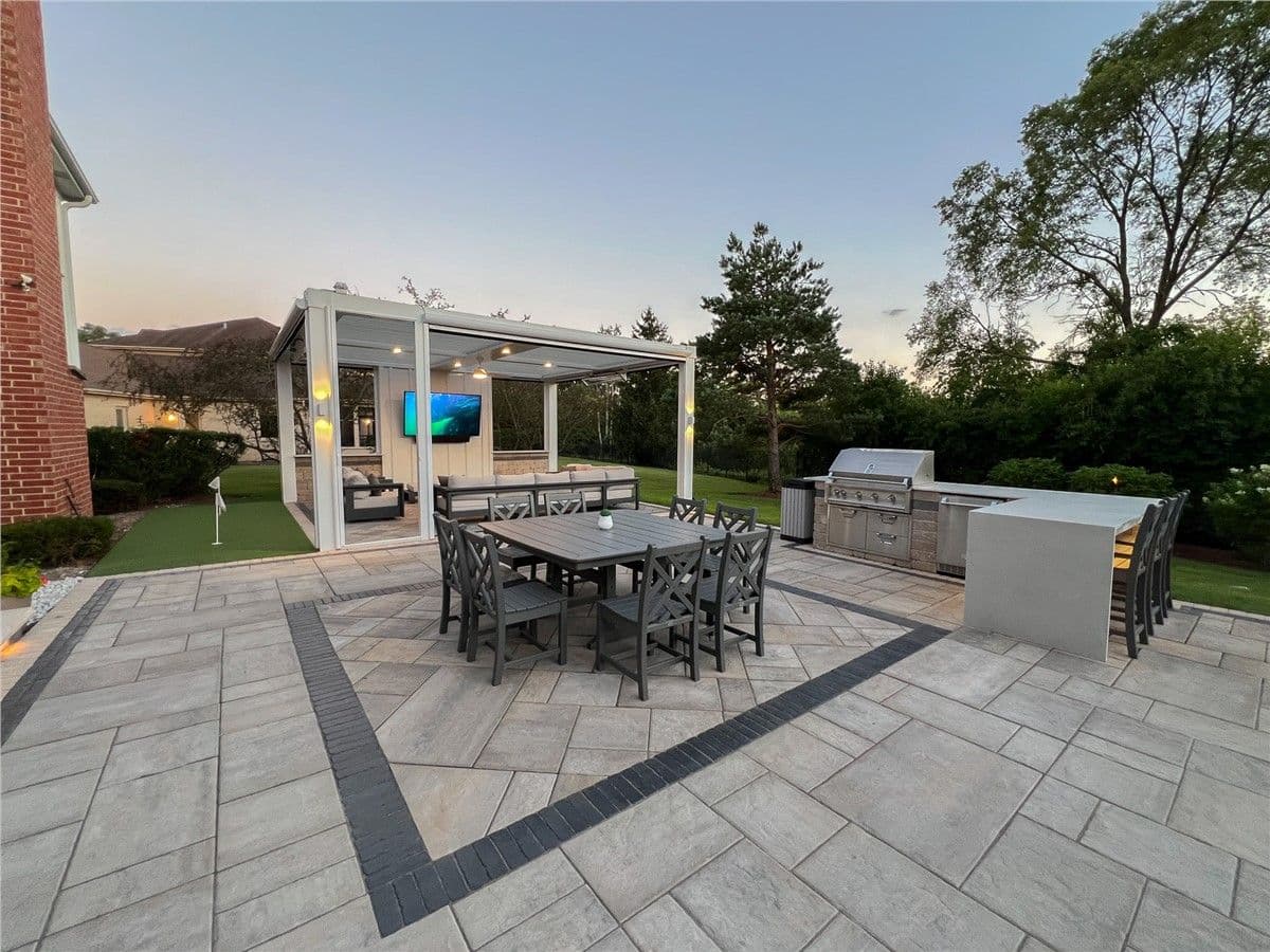A dark outdoor dining table and chairs sit in the center of a stone patio. An outdoor kitchen and grill are off to the right and a white pergola with seating is in the back left.