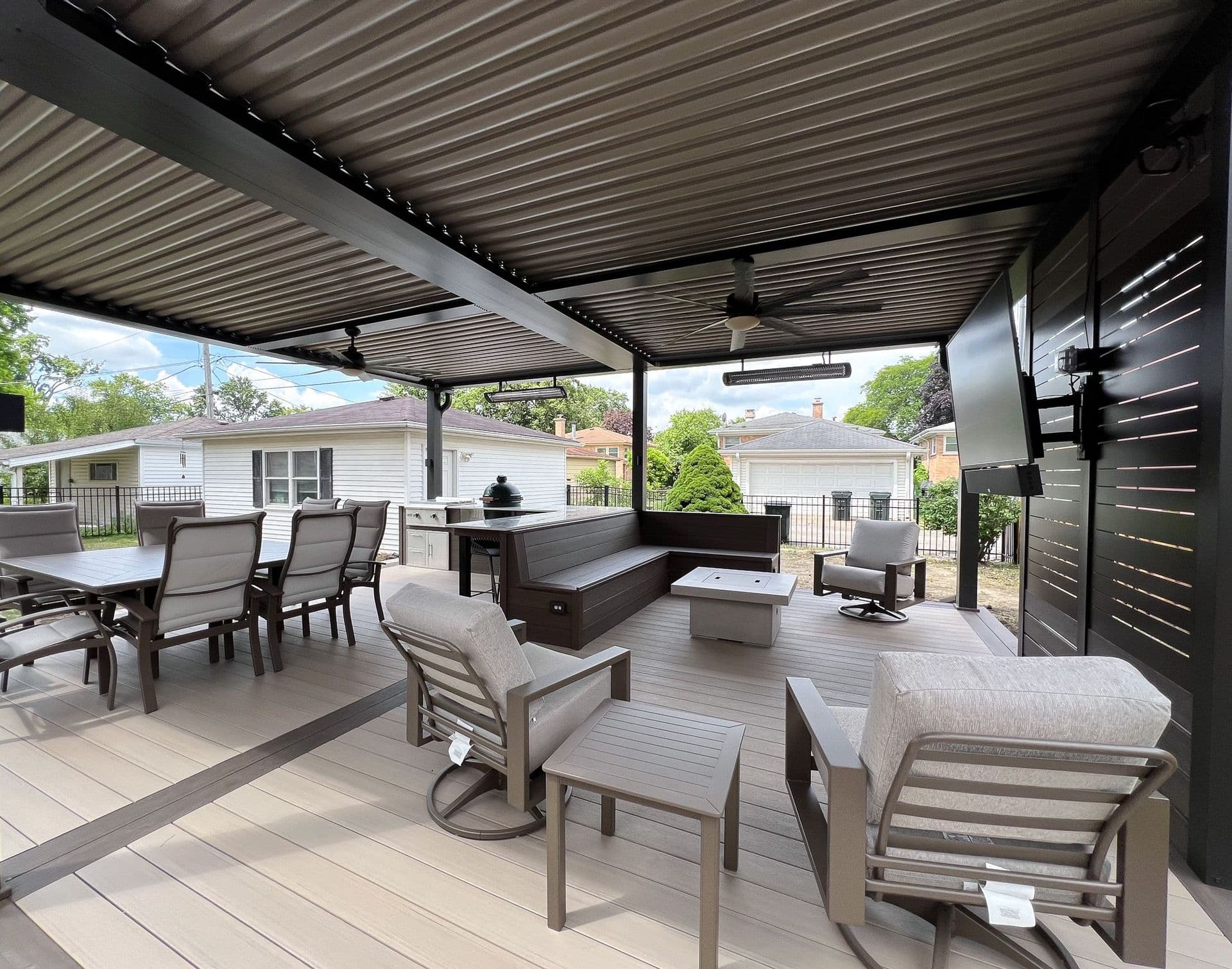 A large black louvered pergola covers light patio furniture and includes features such as built-in fans and a television.