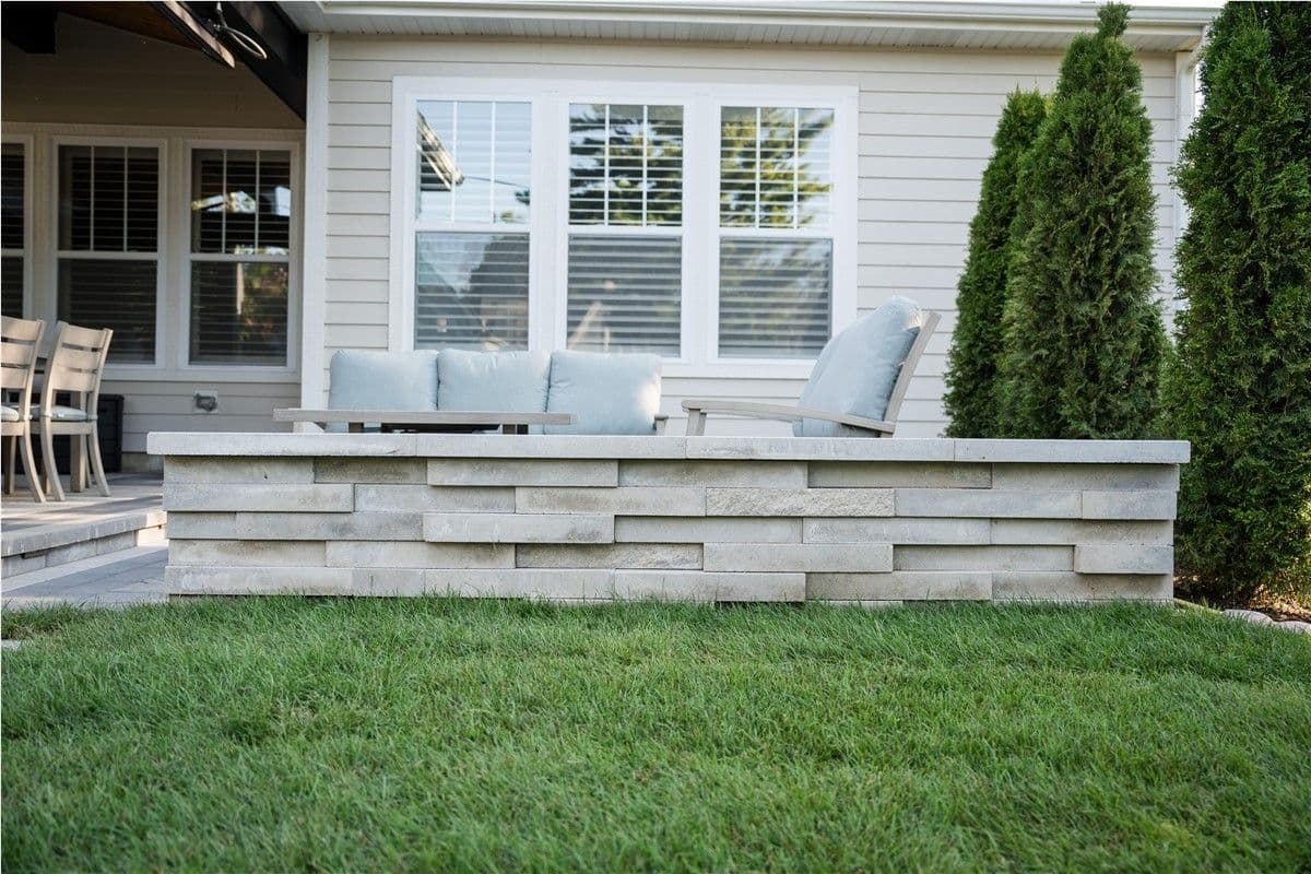 A low stone wall in front of a patio with light-colored patio furniture. Green grass is in the foreground.