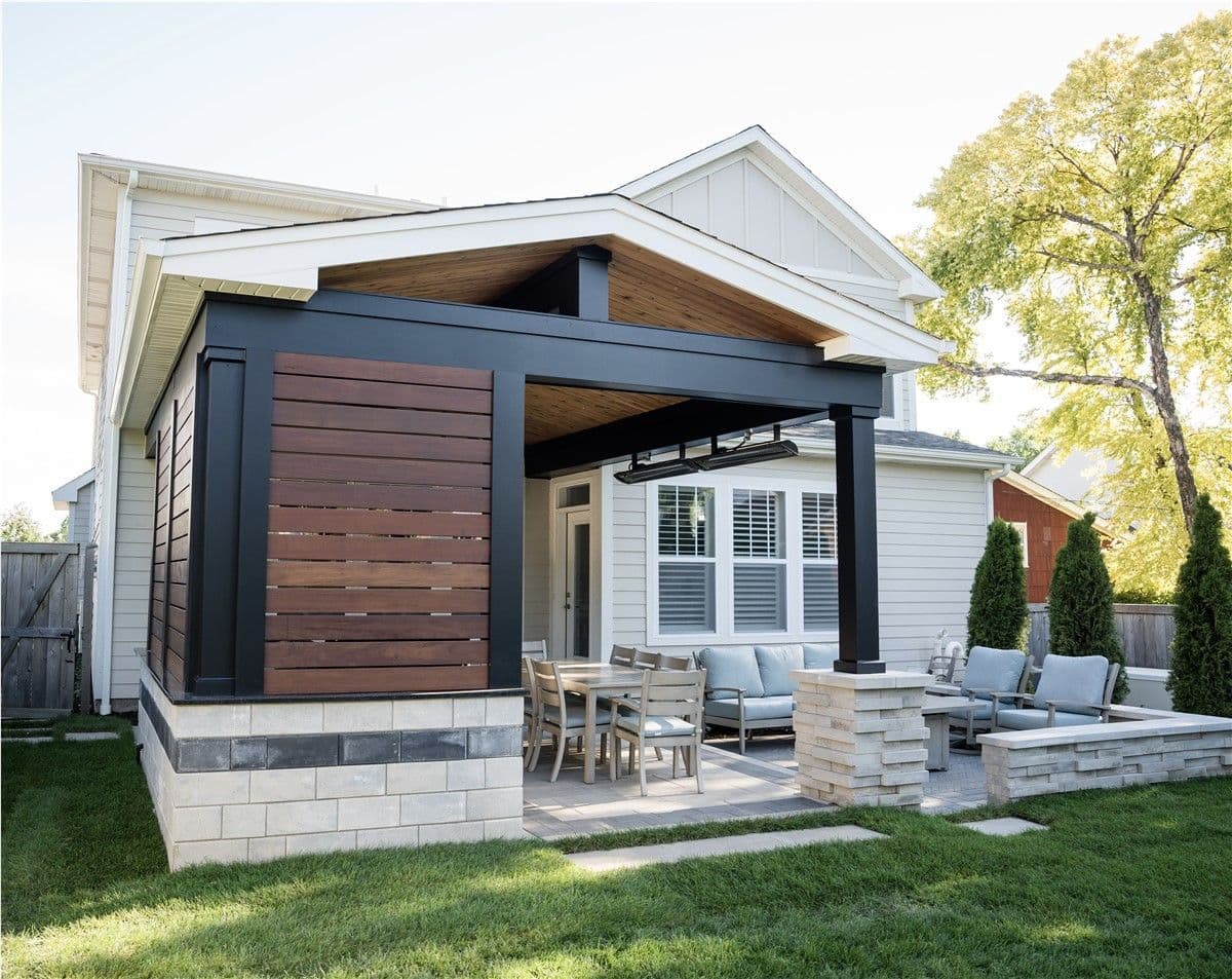 The backyard of a white house with a brown and black covered pergola area and an open area with patio furniture and a fire pit.