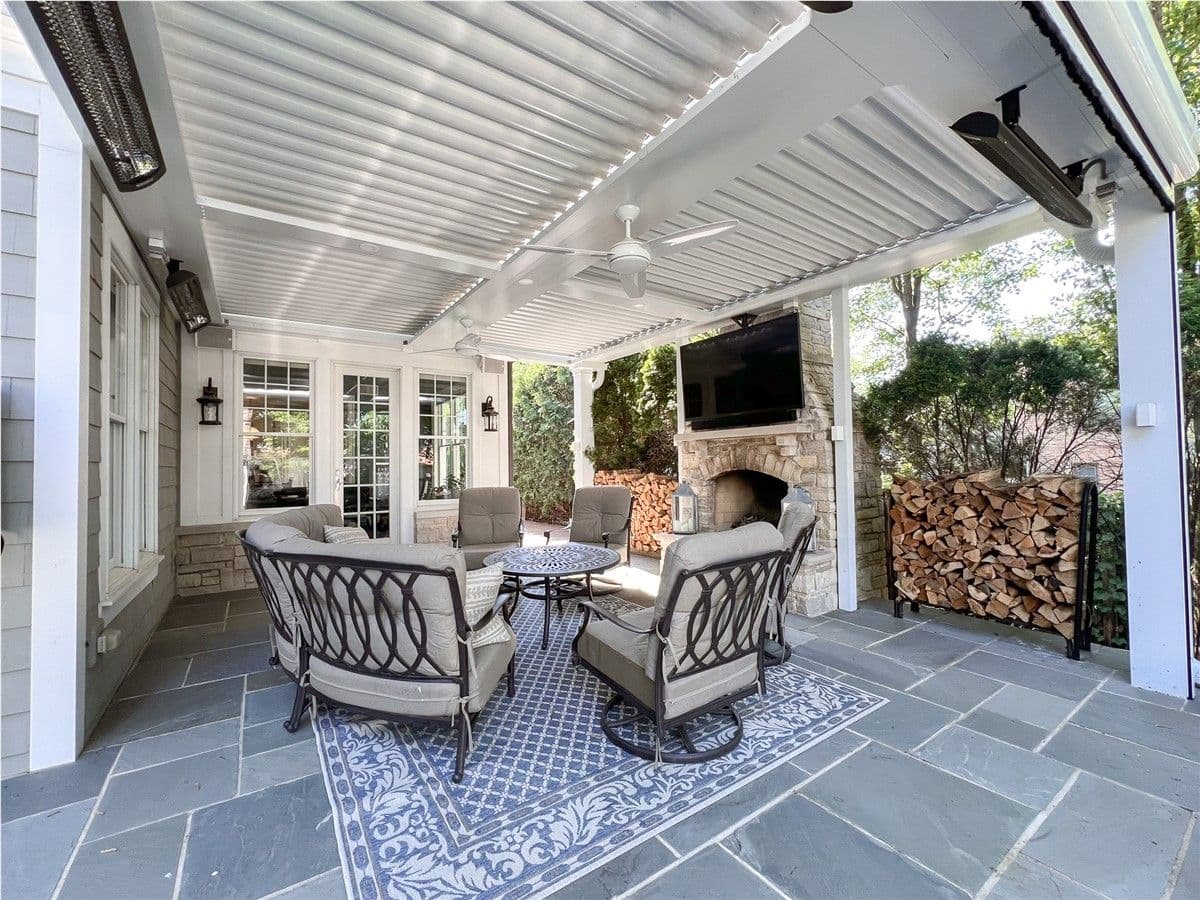 Patio furniture sits in front of a stone fireplace with a mounted TV under a white pergola with louvered roof.
