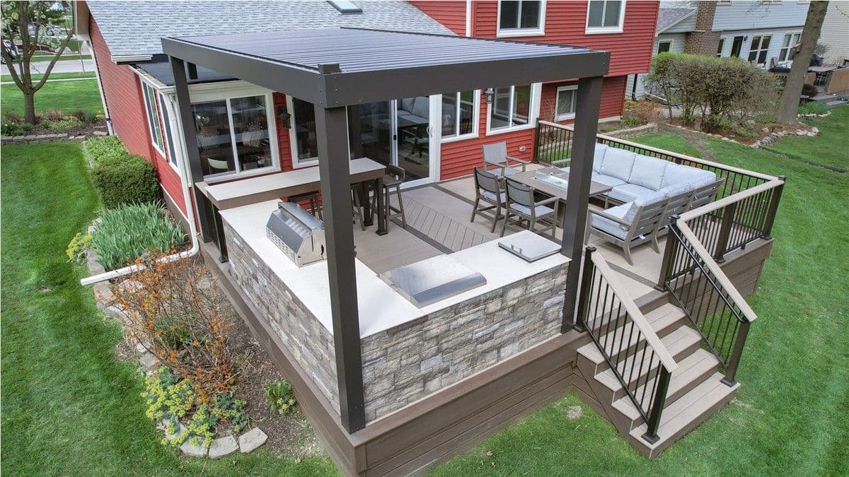Covered outdoor patio attached to a red house, featuring a wood-paneled ceiling with a ceiling fan, light stone flooring, dark wicker seating with beige cushions, and a stone fireplace.