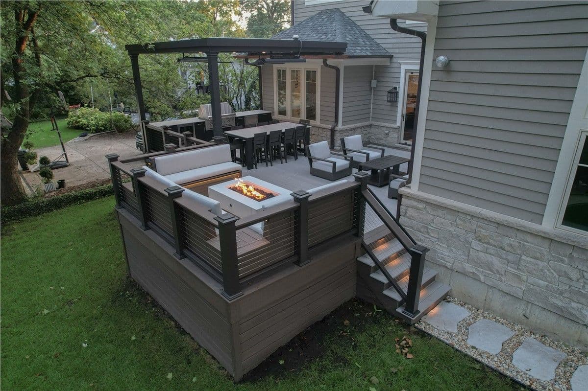 Outdoor living area with a deck, pergola, built-in grill station, fire pit, and surrounding seating, complemented by greenery and a landscaped garden.