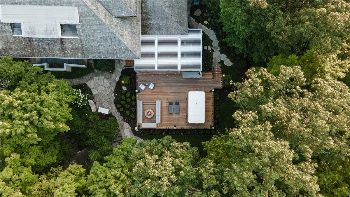 An aerial view of a back deck with a white pergola, bench seating around a fire pit, and lounge chairs.