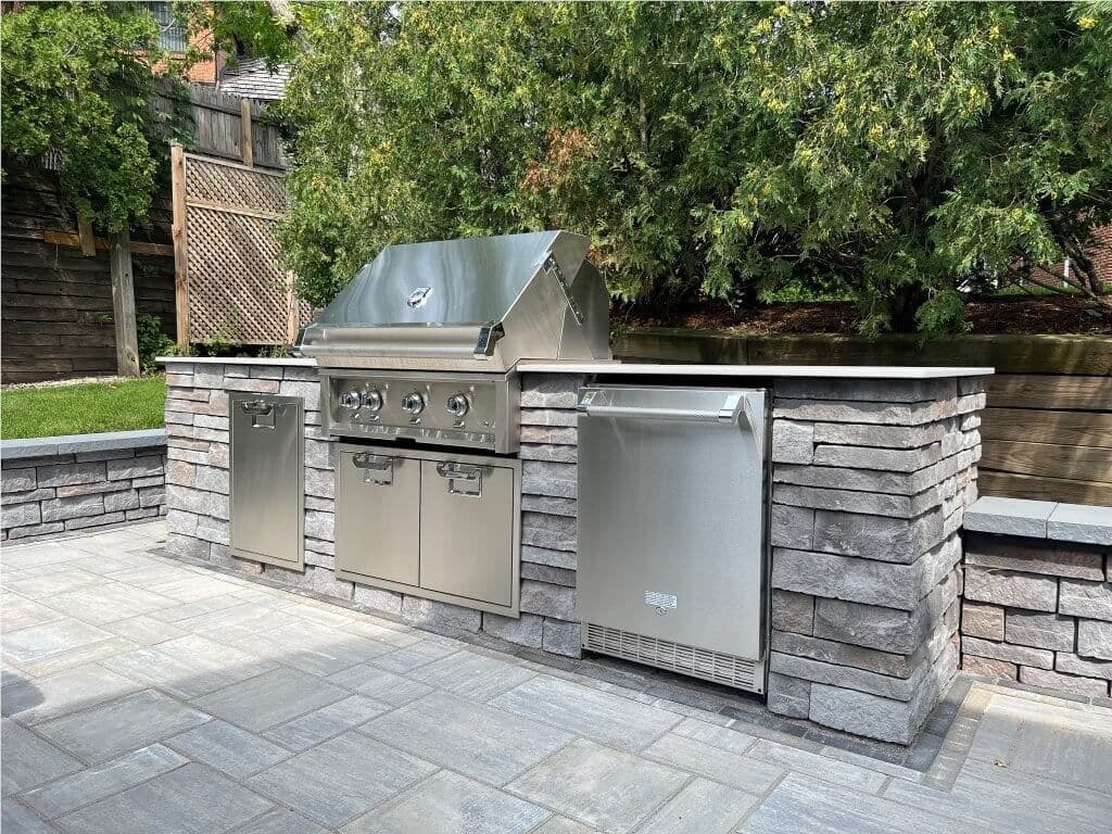Outdoor kitchen with a built-in stainless steel grill set in a stone island on a paver patioin a yard surrounded by trees and shrubs.