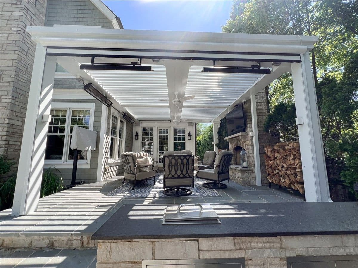 A patio with furniture, stone fireplace, and TV underneath a white pergola with an open louvered roof.