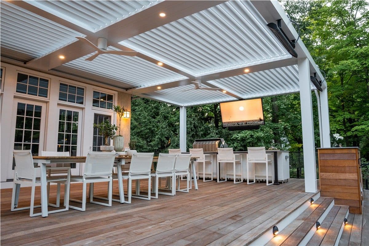 A deck with a long dining table and white chairs towards the left, and bar seating with white bar stools towards the back right under a white pergola. A grill is visible in the background.