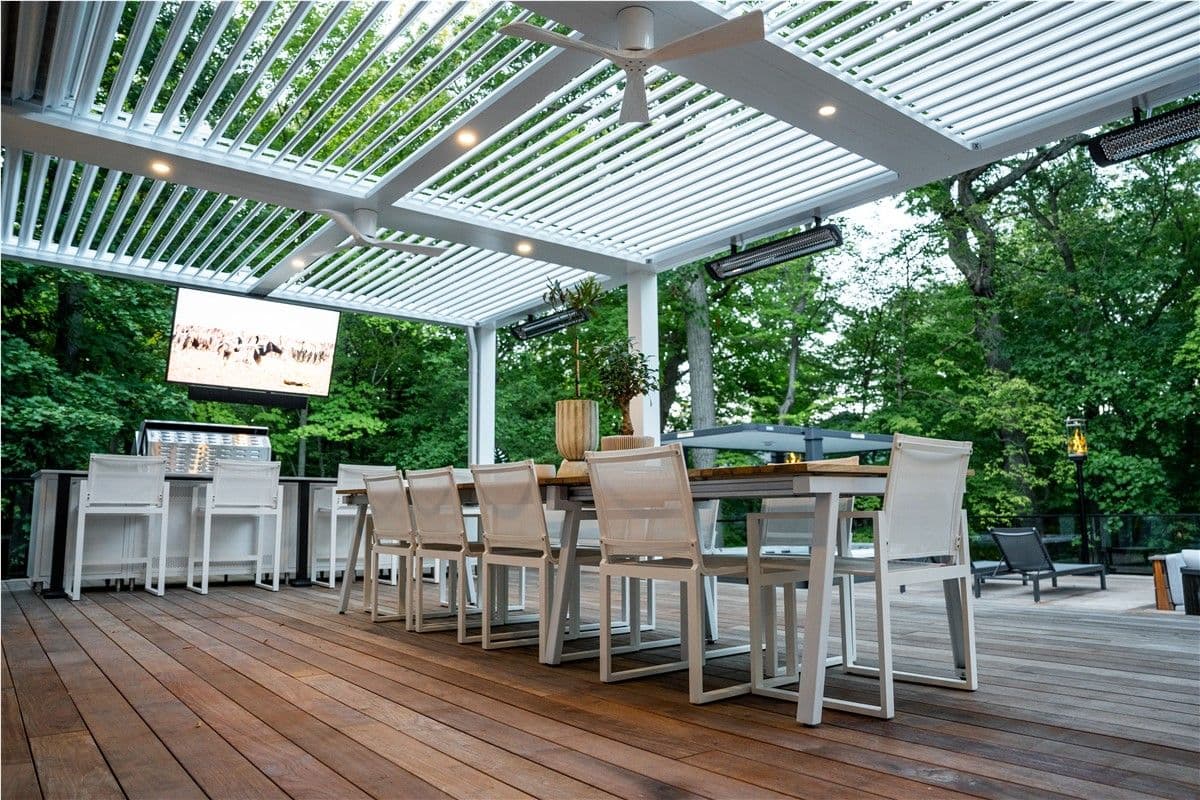 A deck with a long dining table and white chairs towards the left, and bar seating with white bar stools towards the back right under a white pergola. A grill is visible in the background.