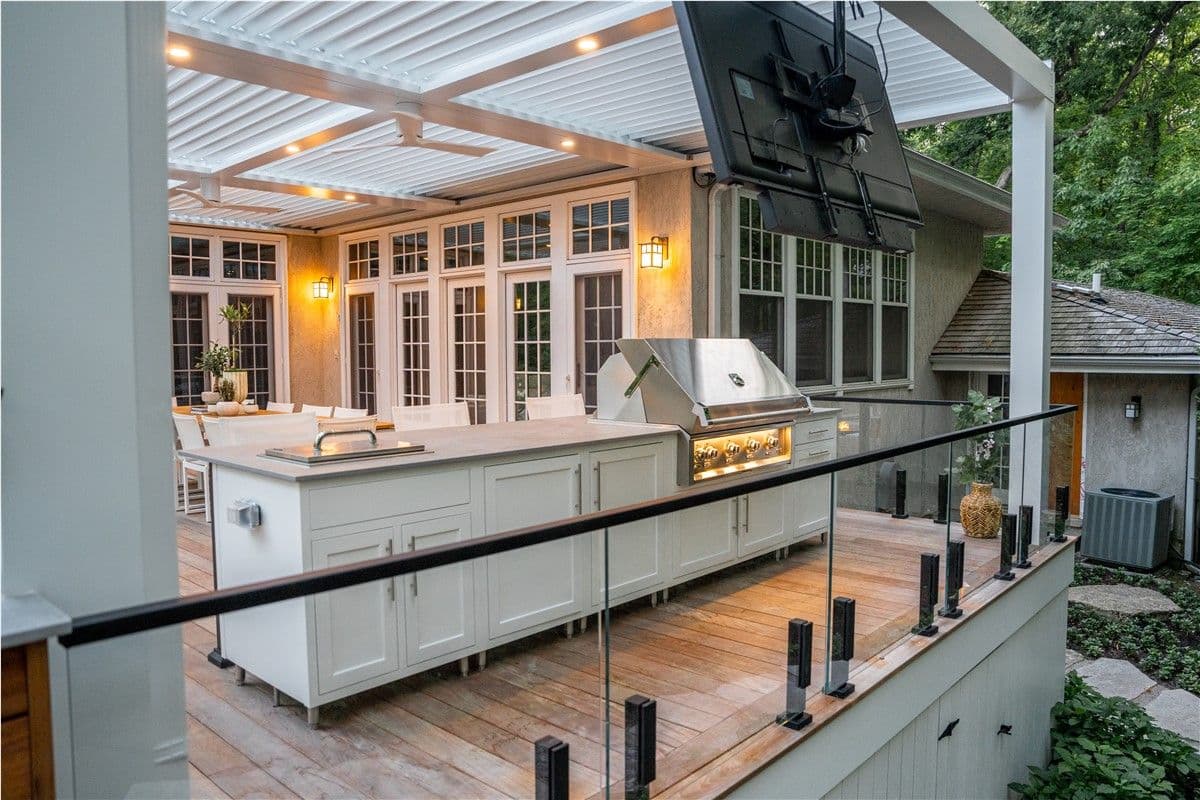 A view of a deck with an outdoor kitchen area with white cabinets and built-in grilling station. A TV is mounted from a white pergola.