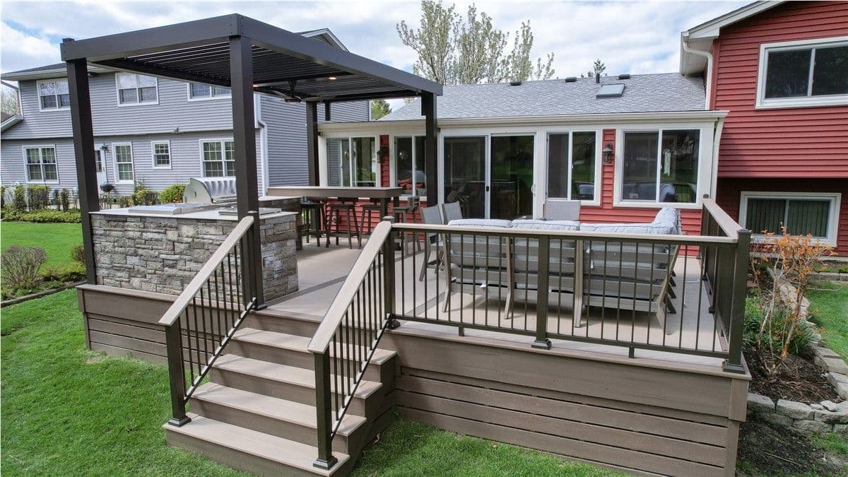 A light brown deck off the back of a red and white house. There is a black pergola with a stone wall, built-in grill, and bar seating on the left, and patio chairs and couch on the right. Steps lead down to the yard in the center.