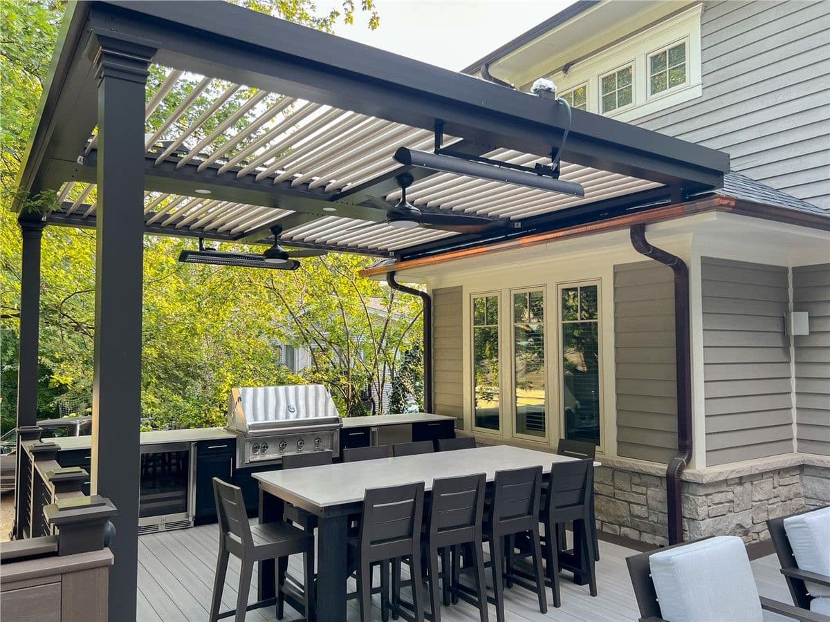 A black and gray deck with a large dining area and chairs. A black pergola with a fan and some heating lamps hangs above, and a built-in grill and kitchen area is in the background.