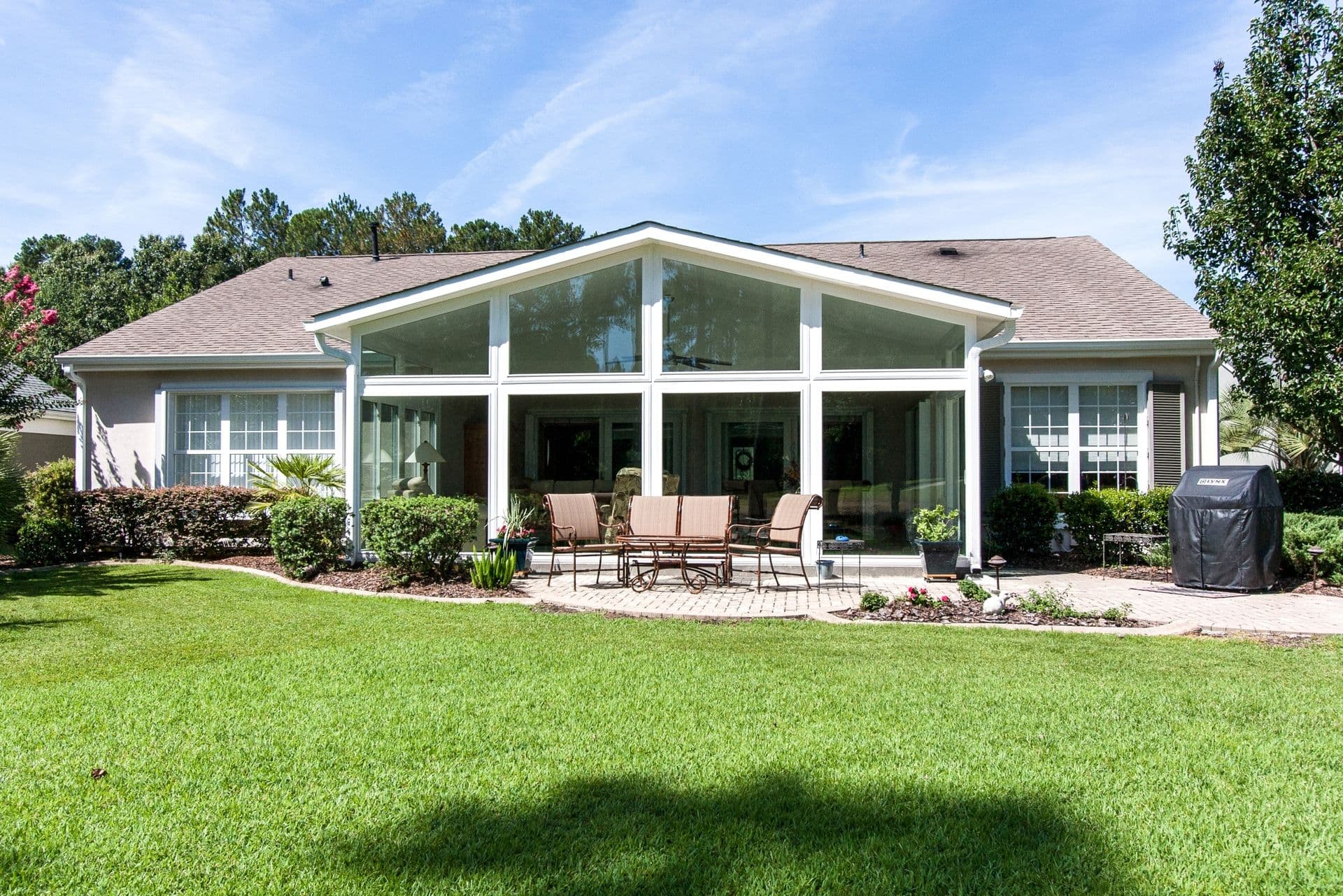 Sunroom with white-framed windows and a gable roof, attached to a light-colored house and surrounded by a lawn and landscaped garden beds.