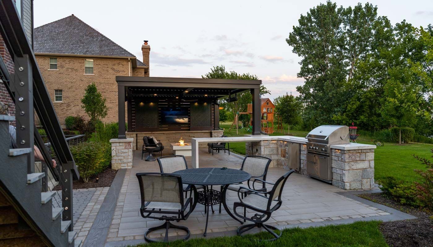 Spacious backyard with a gray paver patio, dark wicker outdoor furniture with light cushions, and a wooden pergola, surrounded by green lawn and mature trees.