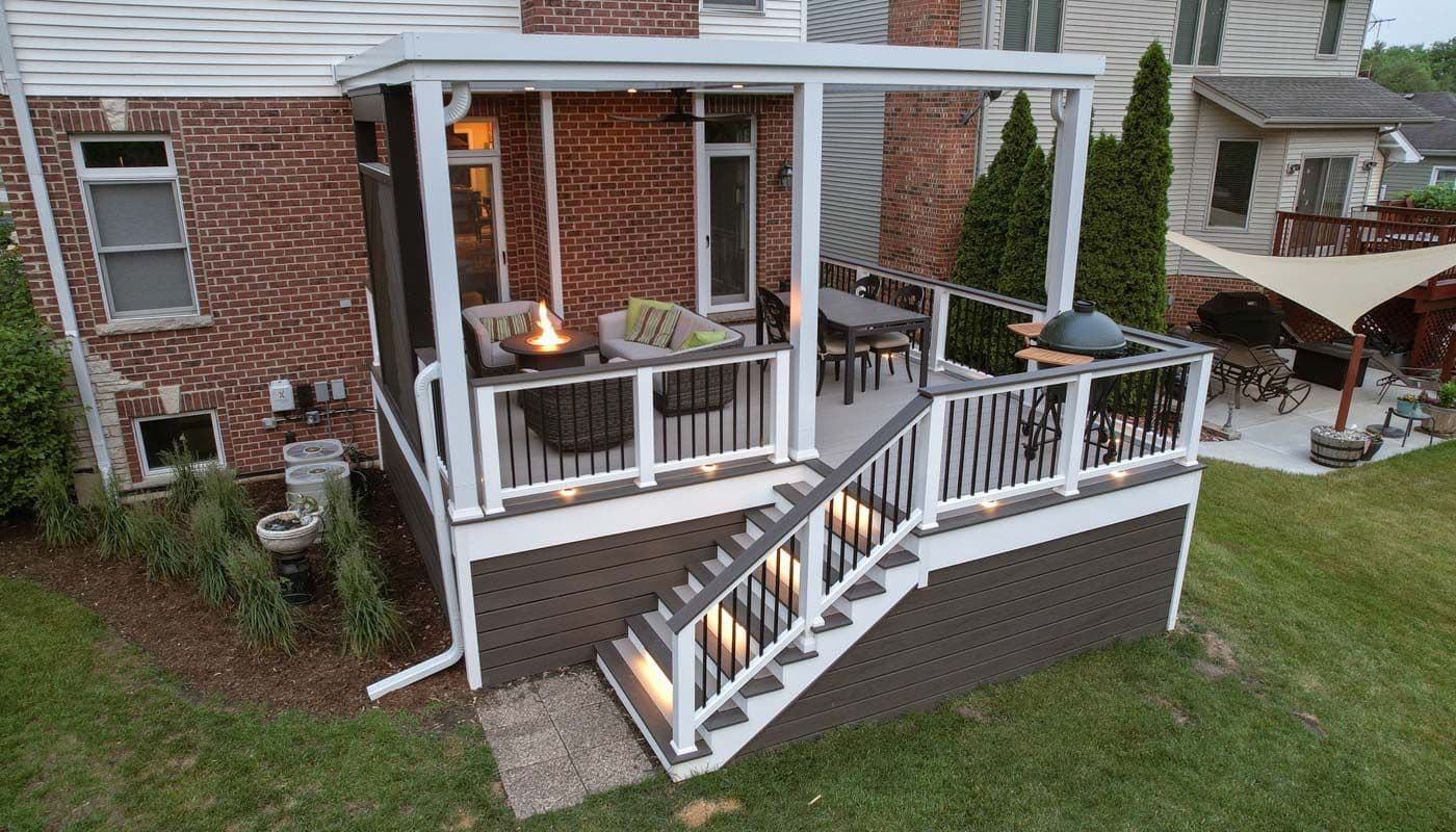 An outdoor deck and living space with a white and gray pergola, small fire pit, comfortable seating, and steps leading down to a yard attached to a red brick house.
