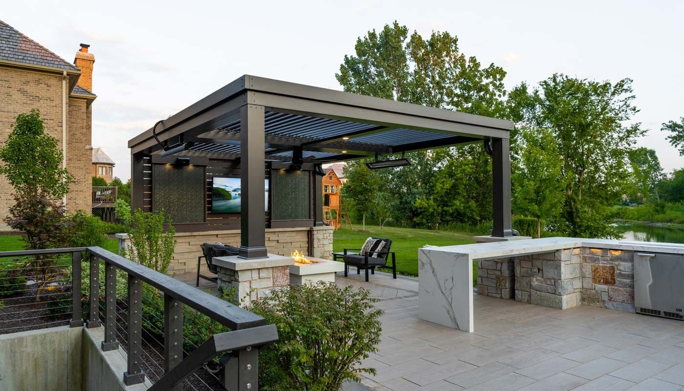 A backyard patio with a dark brown pergola, cushioned outdoor furniture, a fire pit, mounted outdoor TV, and stone pavers, surrounded by shrubs and mature trees.