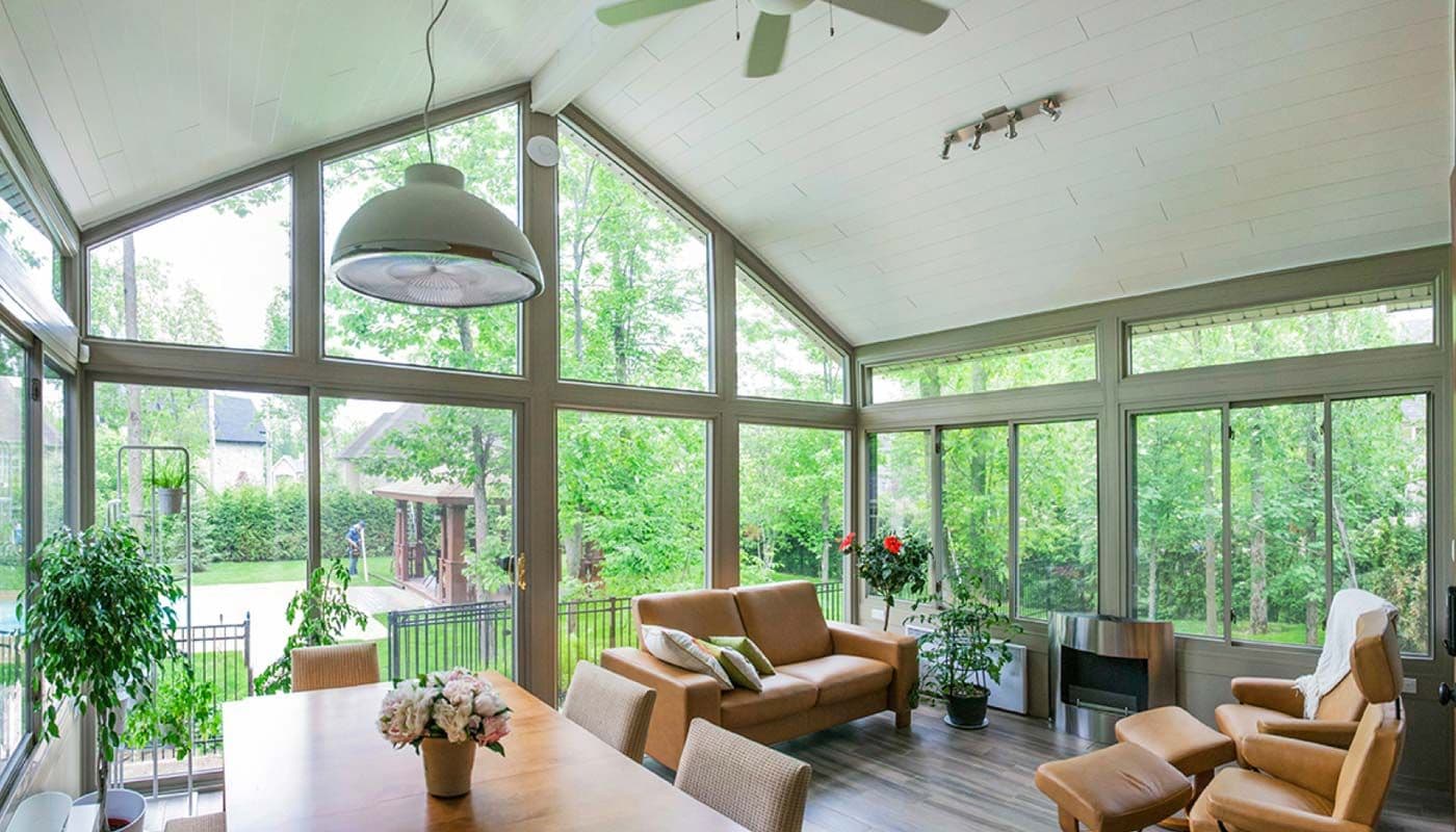 Sunroom with large floor-to-ceiling windows, white framing, and a gable roof, attached to a light-colored house and surrounded by a landscaped yard.
