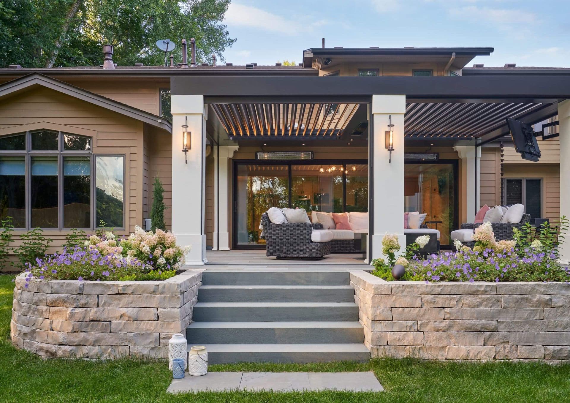 Gray steps between stacked stone garden zones lead to an outdoor patio under a bronze pergola with a mounted TV.