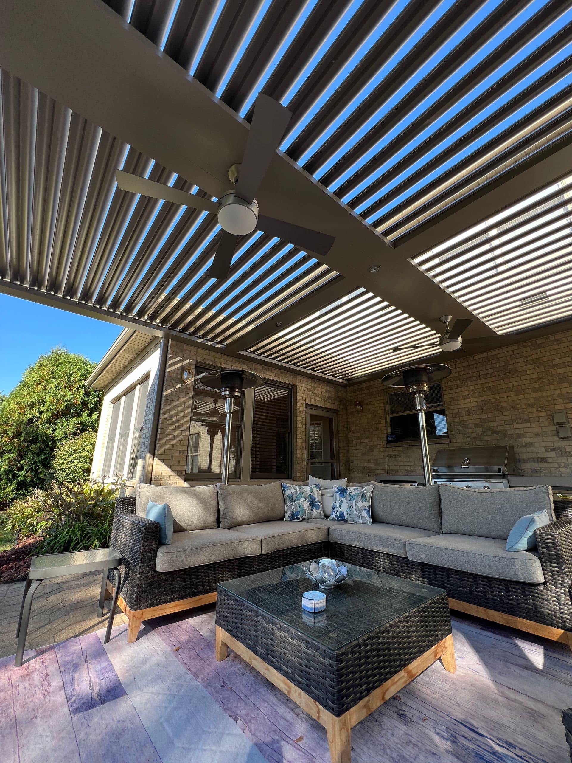 Dark colored wicker patio furniture under a tan louvered roof pergola with hanging outdoor ceiling fans.