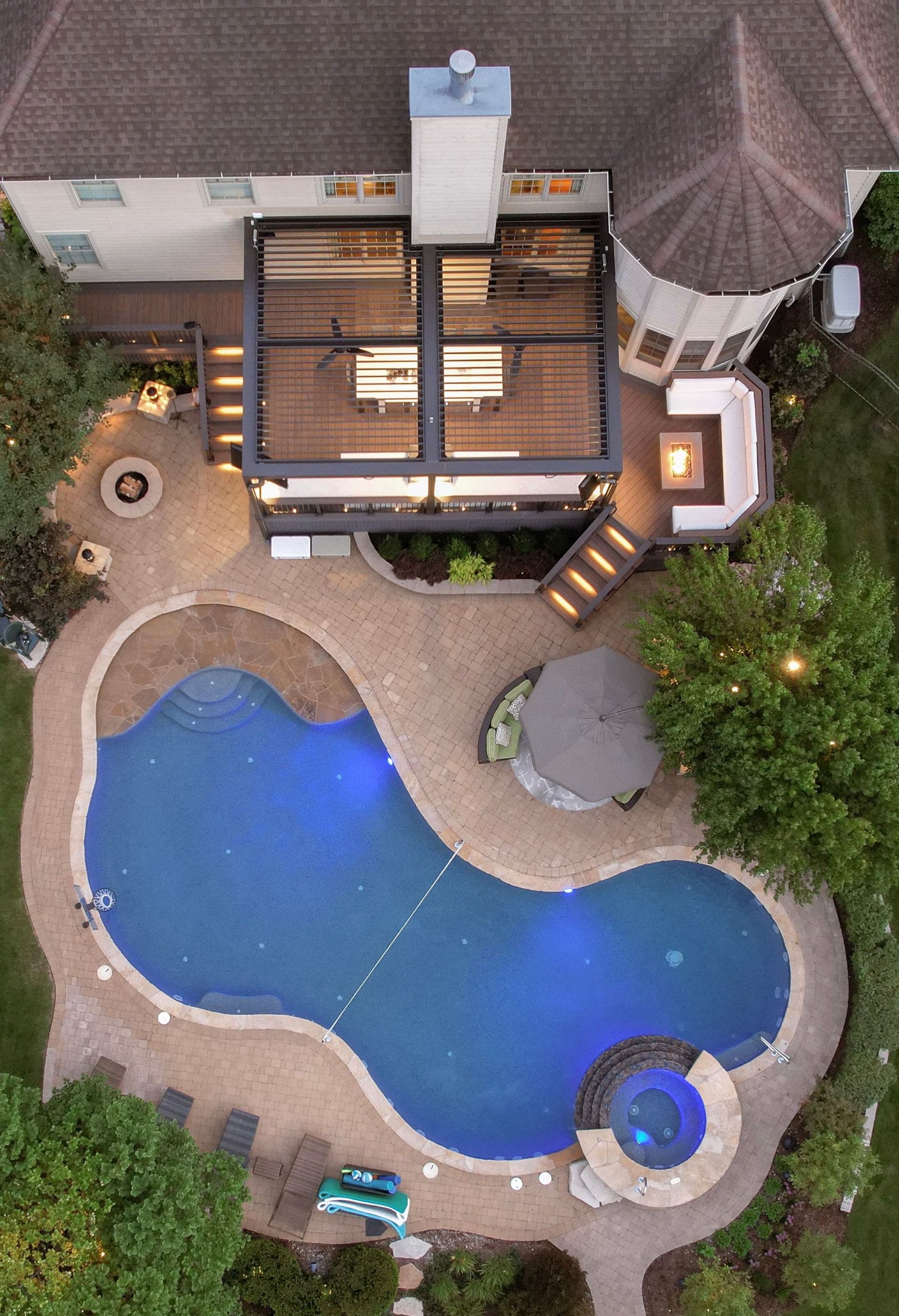 An aerial view of a back deck with a dark-colored pergola. An uncovered seating area with a fire pit is attached to the right. A large pool is in the center surrounded by a patio area.
