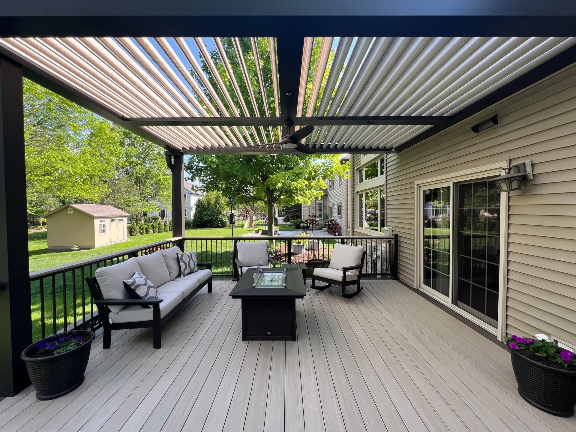 A black custom pergola covers a light colored deck with outdoor patio furniture and a fire pit off of a tan house.
